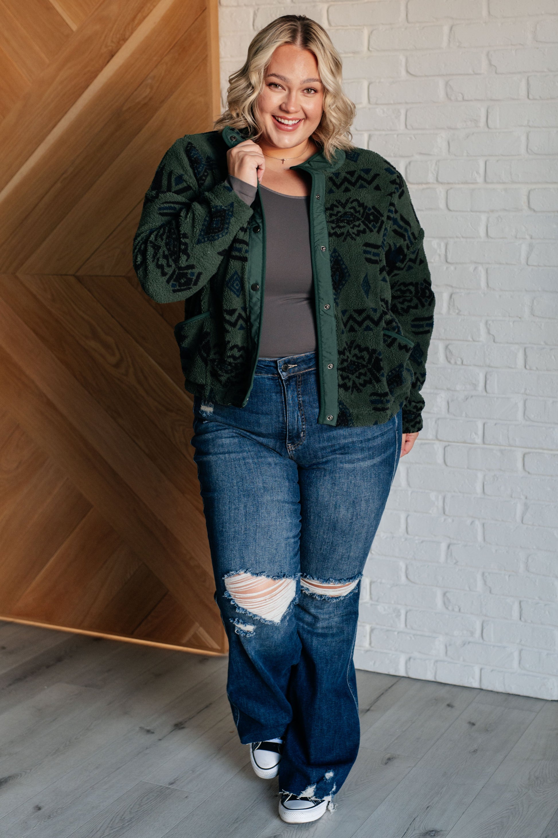 Woman wearing a green patterned jacket, gray top, and blue jeans standing against a white brick wall.