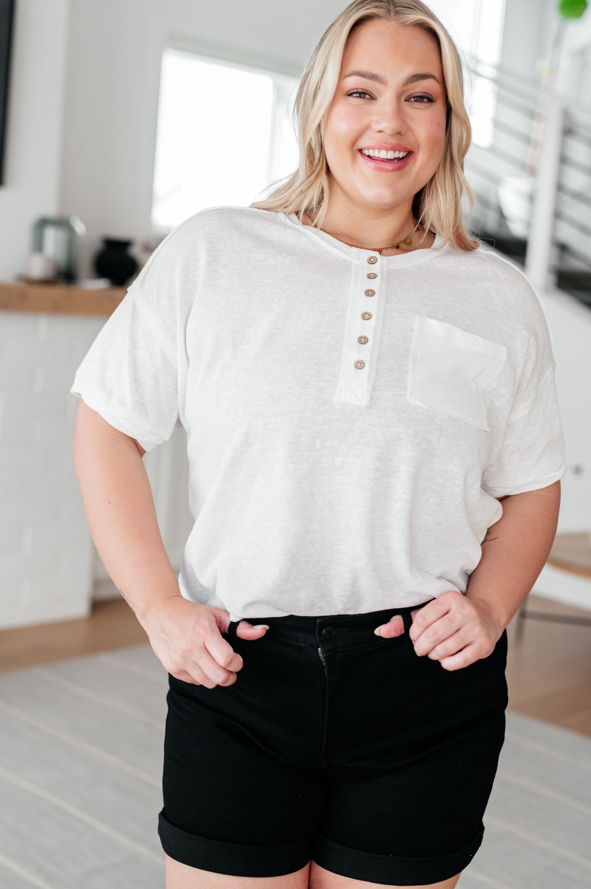 A woman wearing a white Hipster Henley Top with short sleeves, a round neckline, and front button closures. She is also wearing black high-rise shorts.