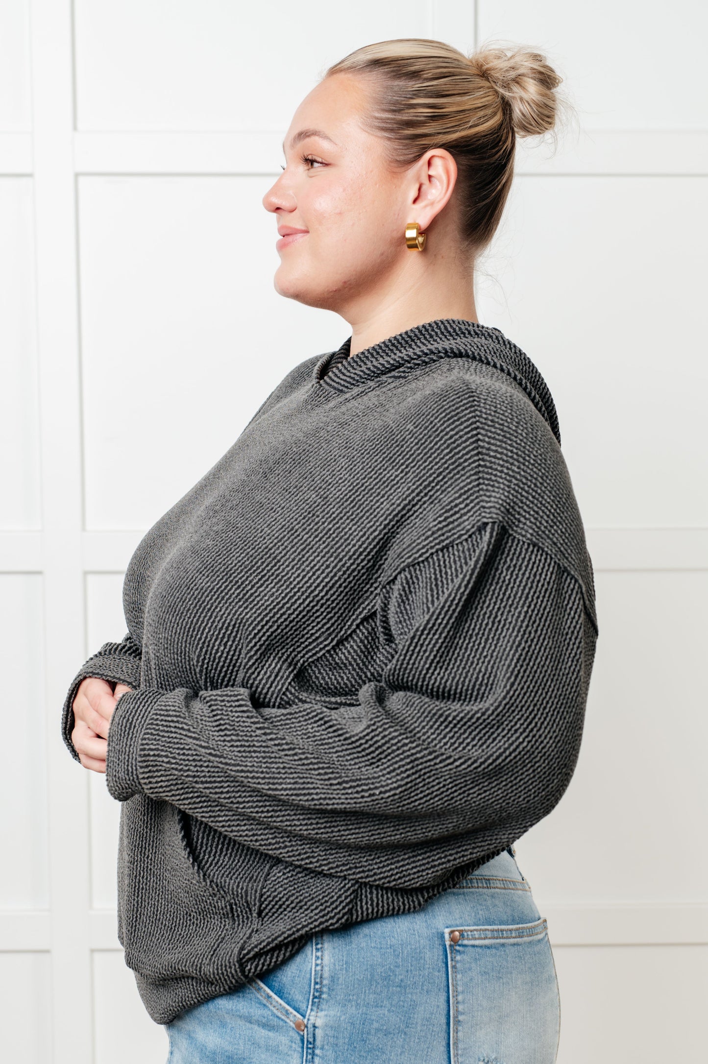 Woman wearing a gray sweater and blue jeans against a white tiled wall.