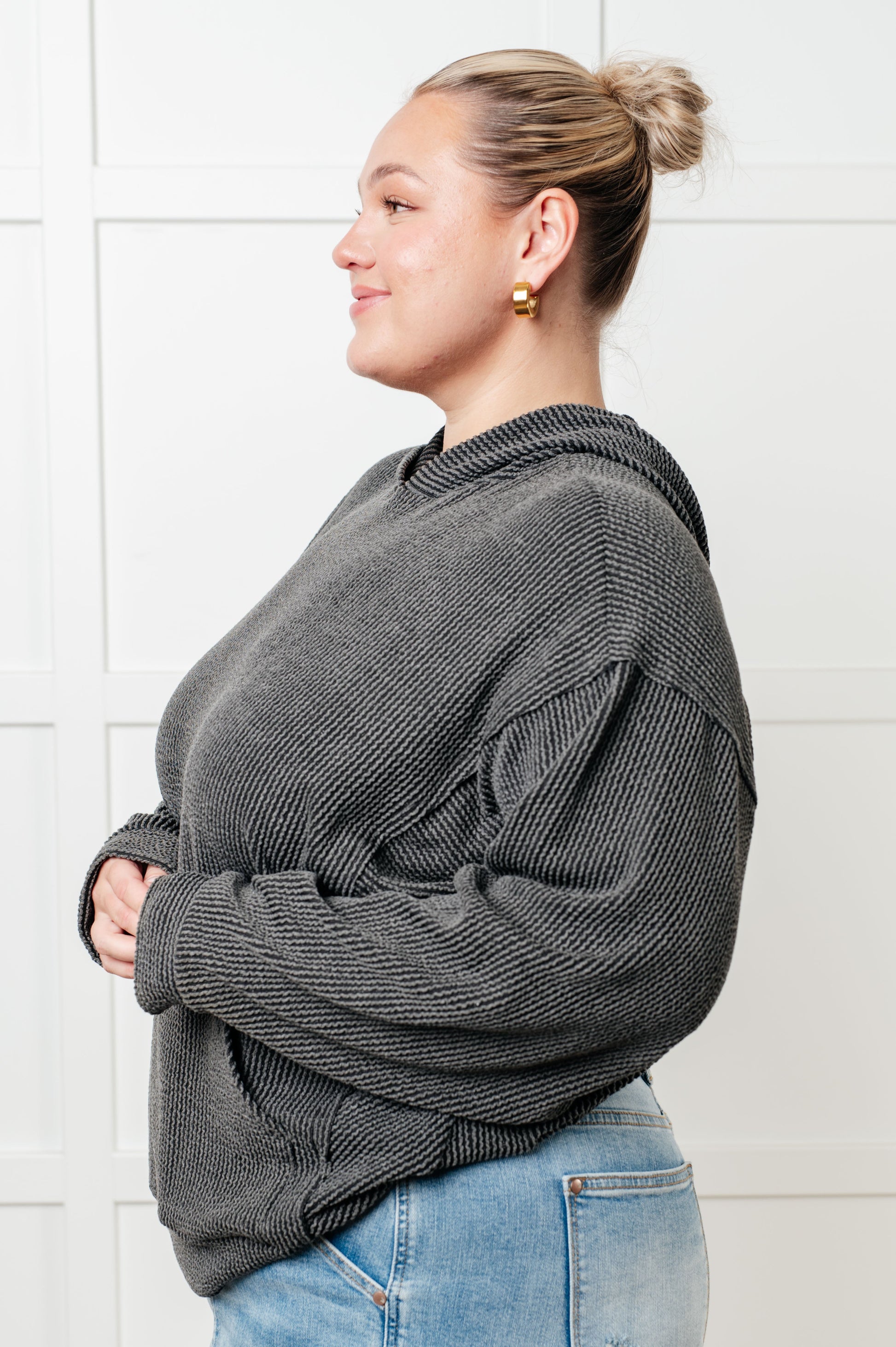 Woman wearing a gray sweater and blue jeans against a white tiled wall.