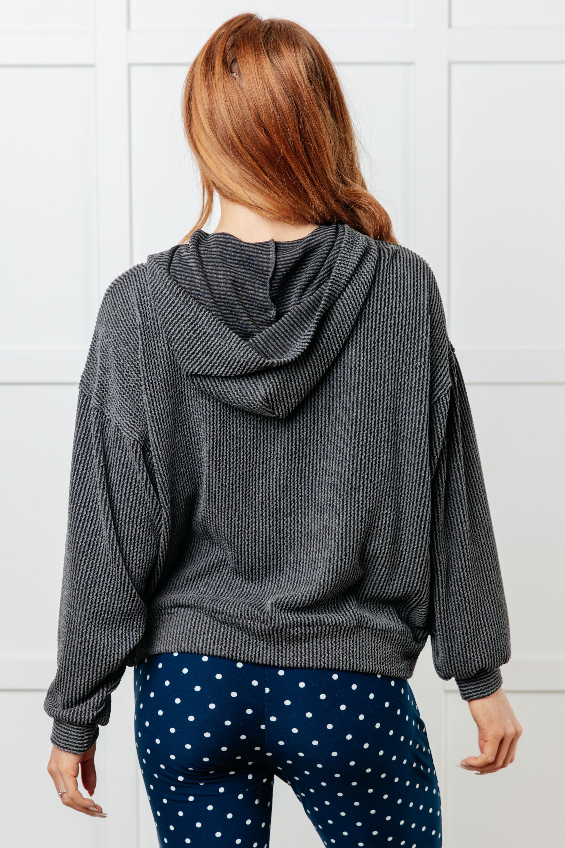 Person wearing a gray hooded sweater and blue polka dot pants against a white tiled wall.