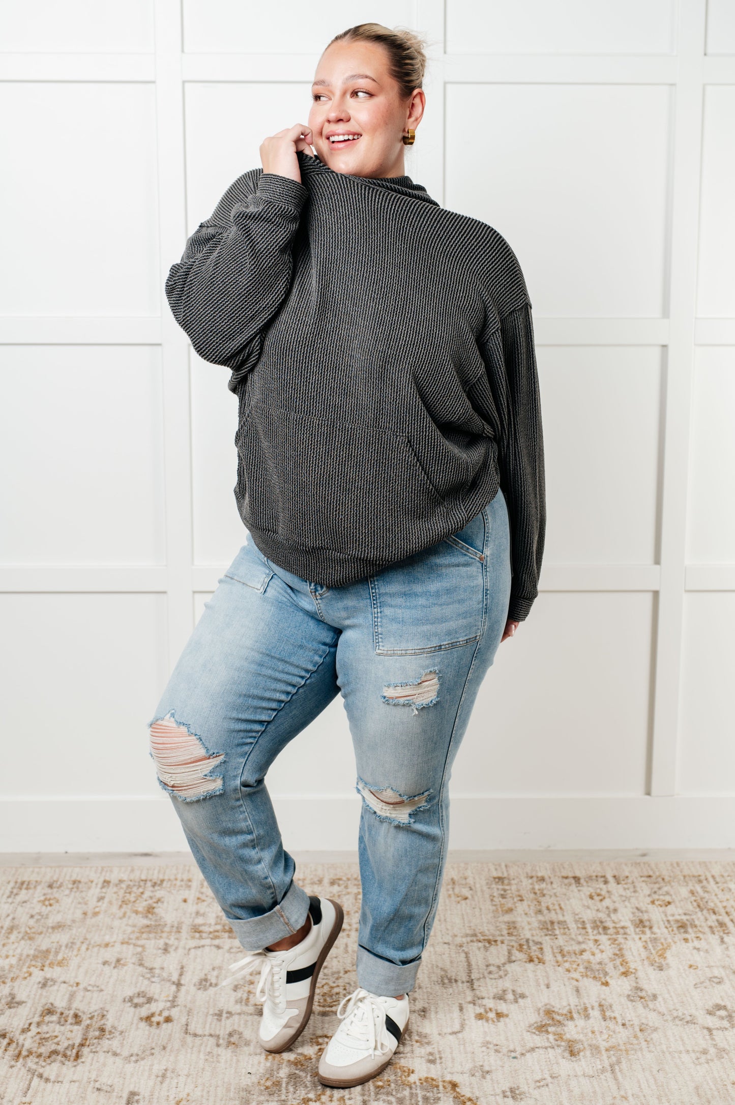 Person wearing a dark gray sweater and light blue jeans standing against a white wall.