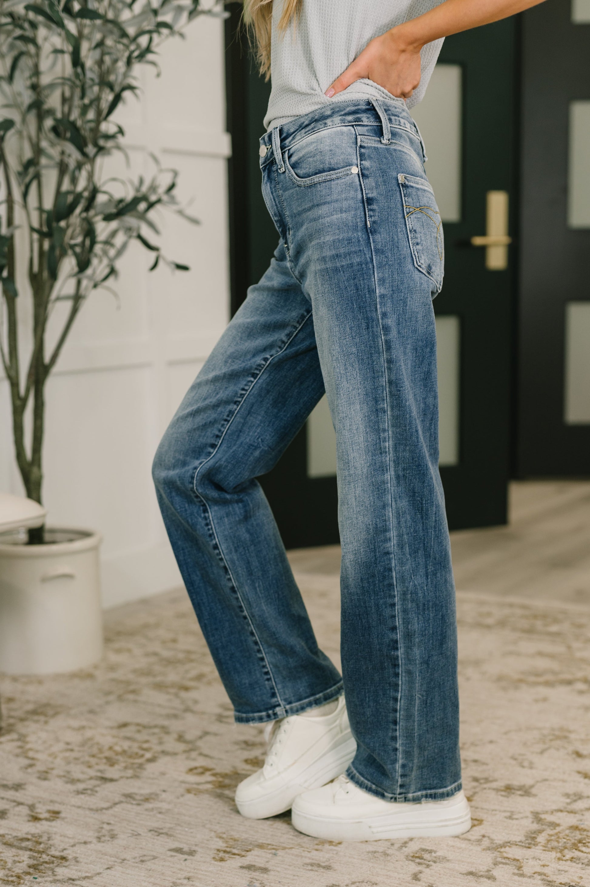 Person wearing blue jeans and white sneakers indoors with a plant and door in the background