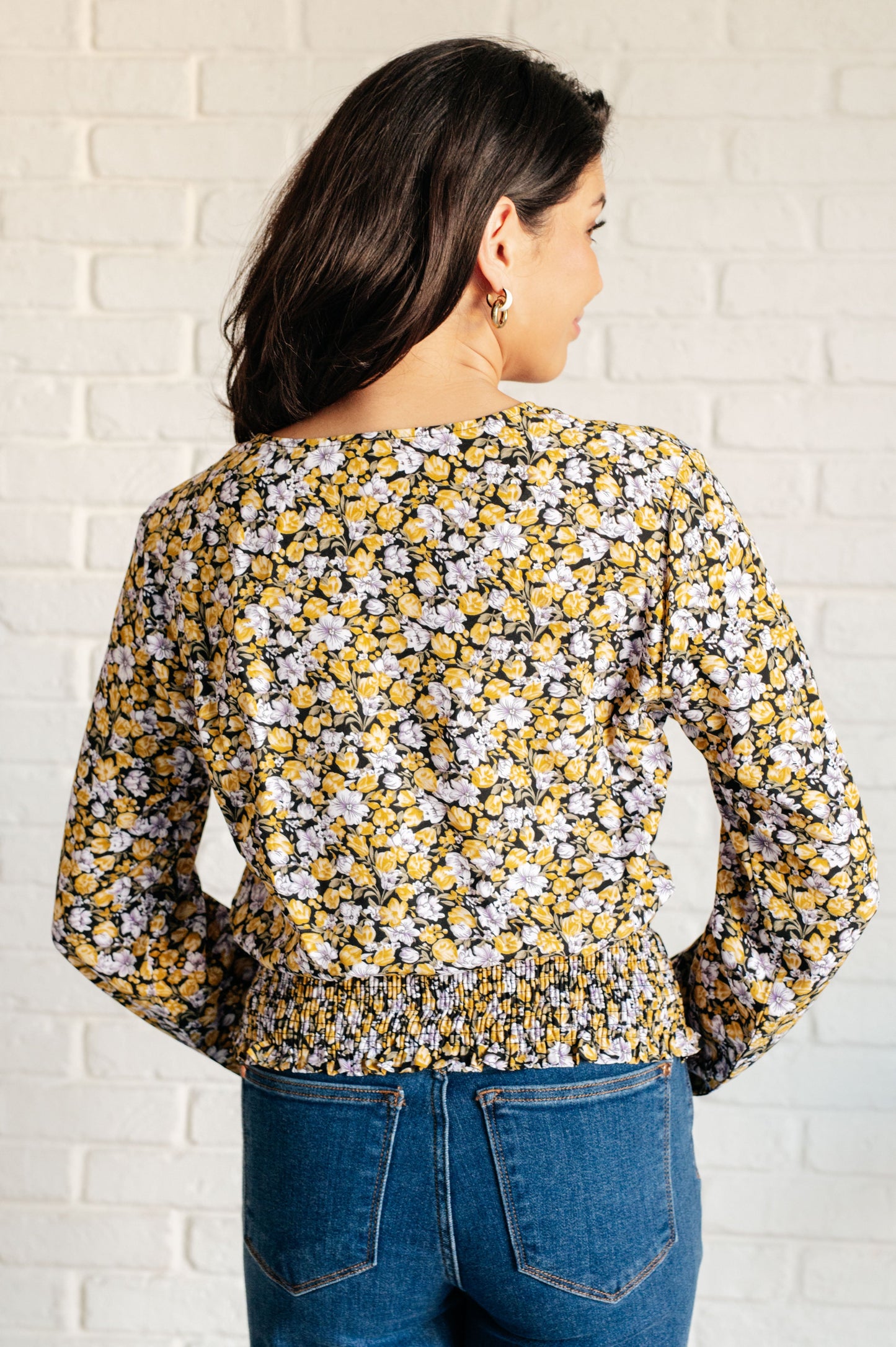 Woman wearing a floral blouse with long sleeves against a white brick wall.