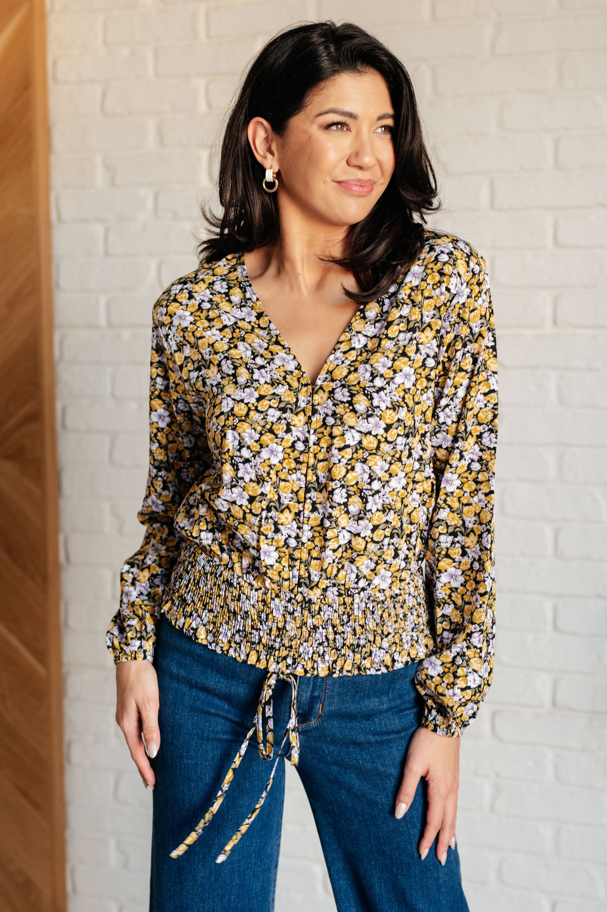 Woman wearing a floral blouse and blue jeans against a white brick wall.