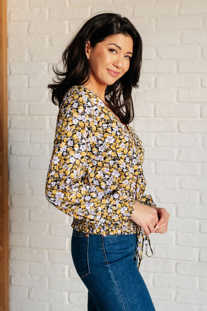 Woman wearing a floral blouse and blue jeans against a white brick wall.