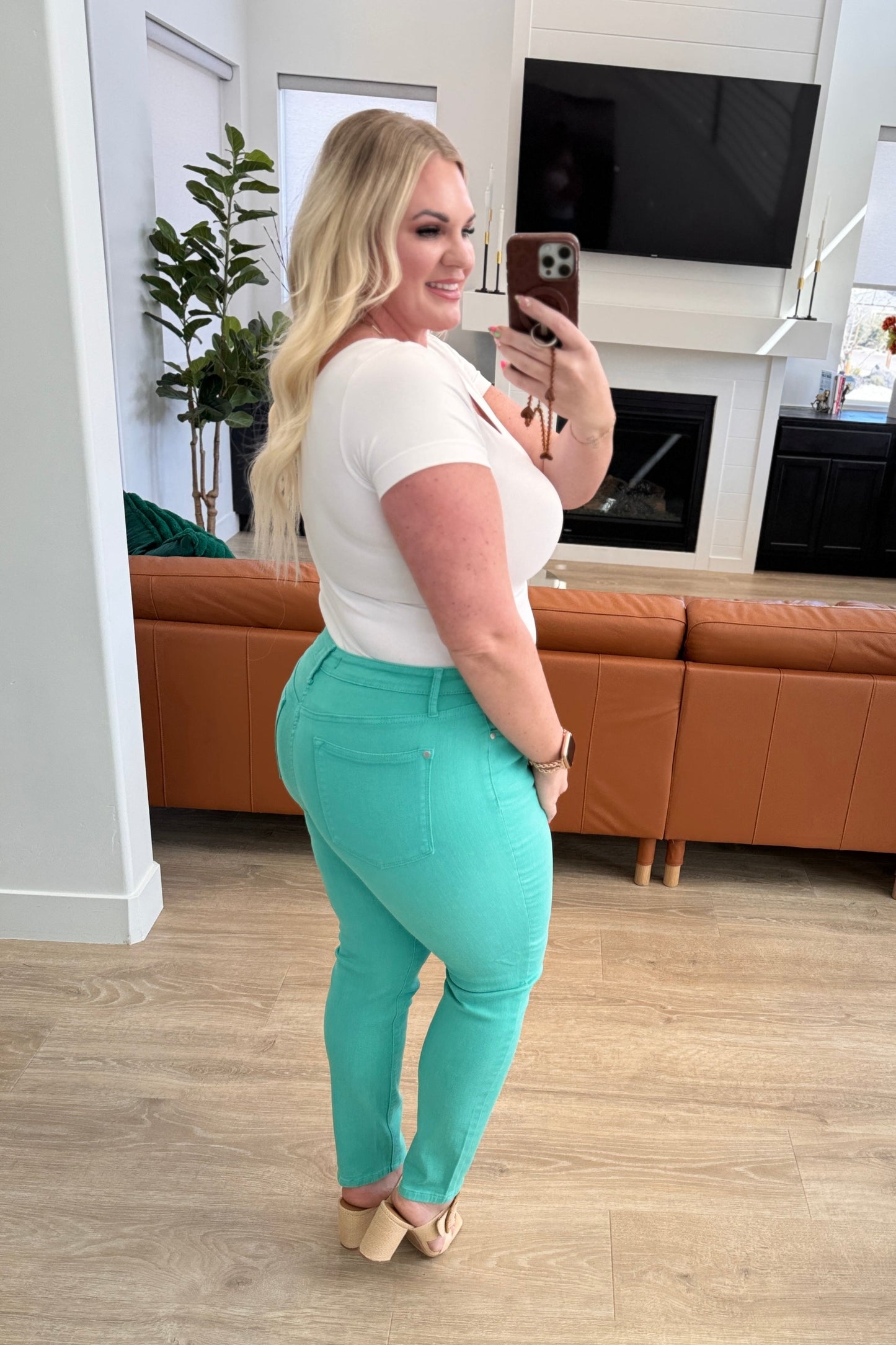 Woman taking a mirror selfie wearing a white top and teal pants in a living room.