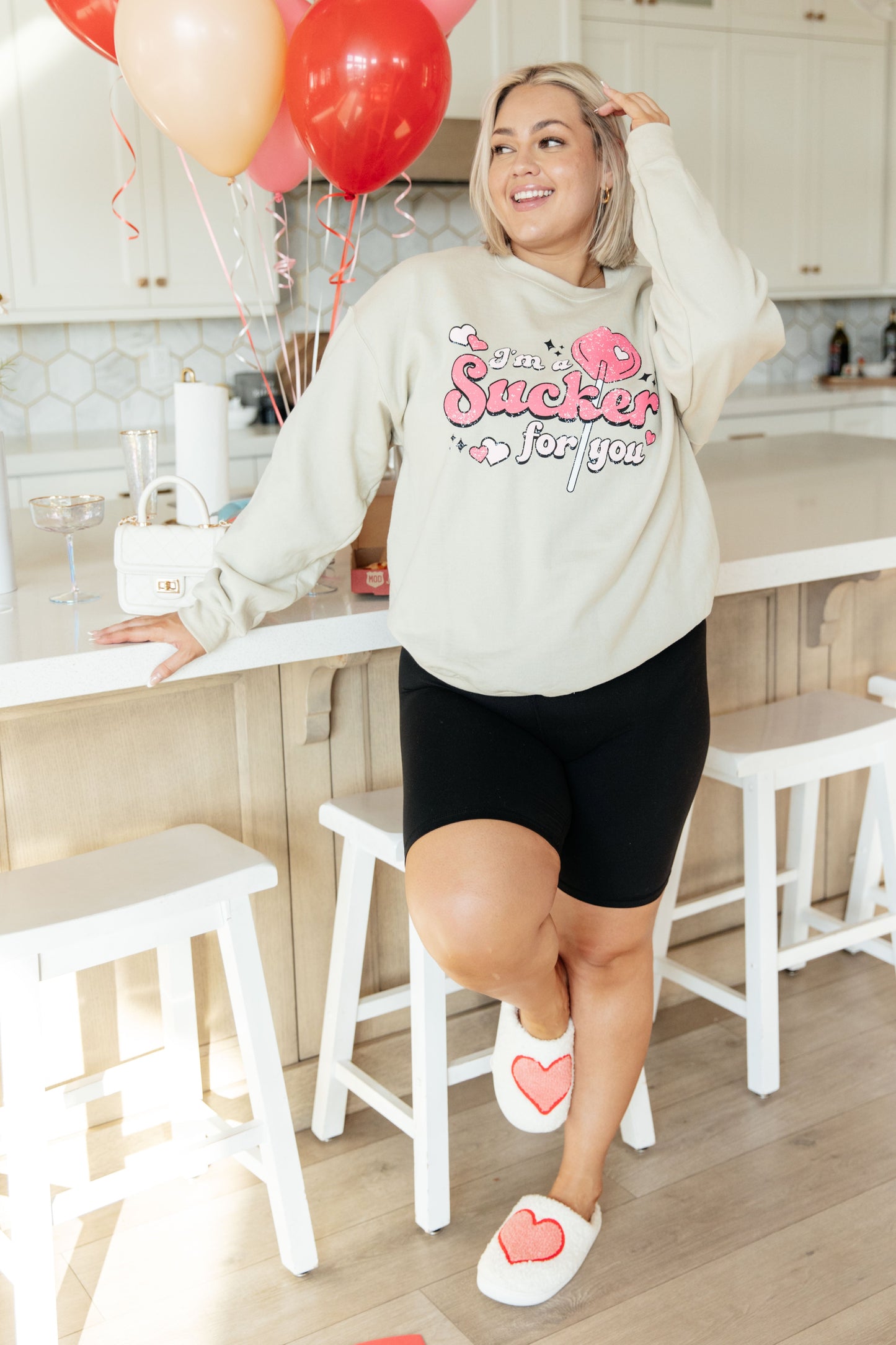Woman in a kitchen wearing a sweatshirt with a heart design, standing near balloons.