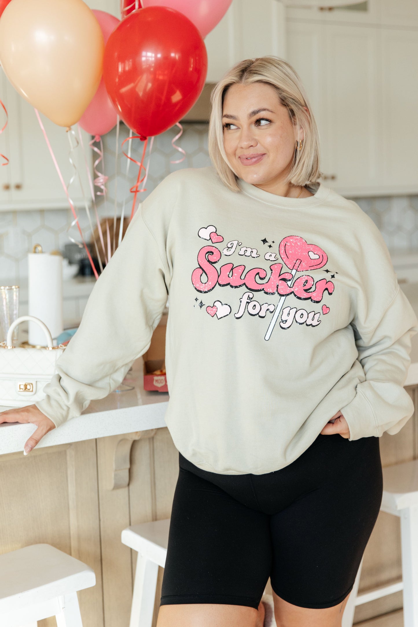 Woman wearing a light gray sweatshirt with a humorous design, standing in a kitchen with balloons in the background.