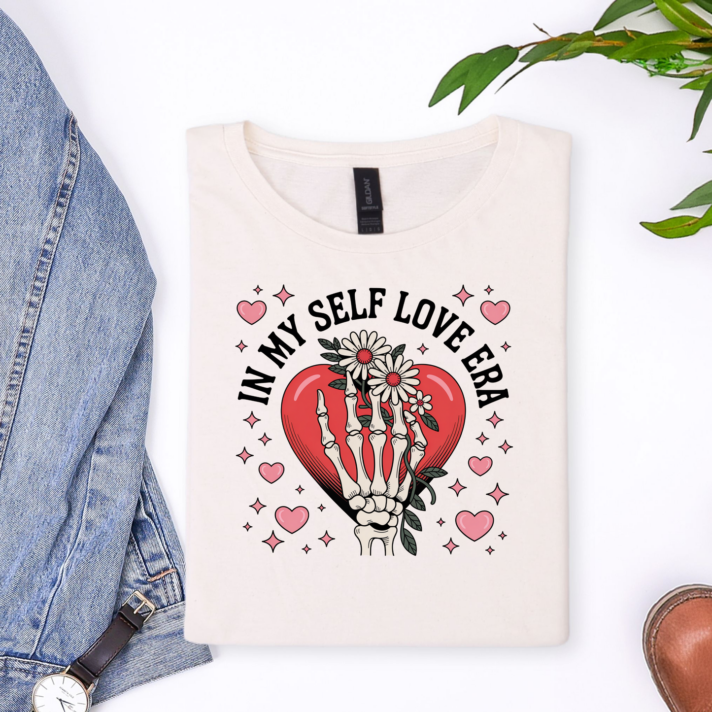 White sweatshirt with skeleton hands holding a heart and 'In My Self Love Era' text, placed on a white surface with denim jeans and a watch.