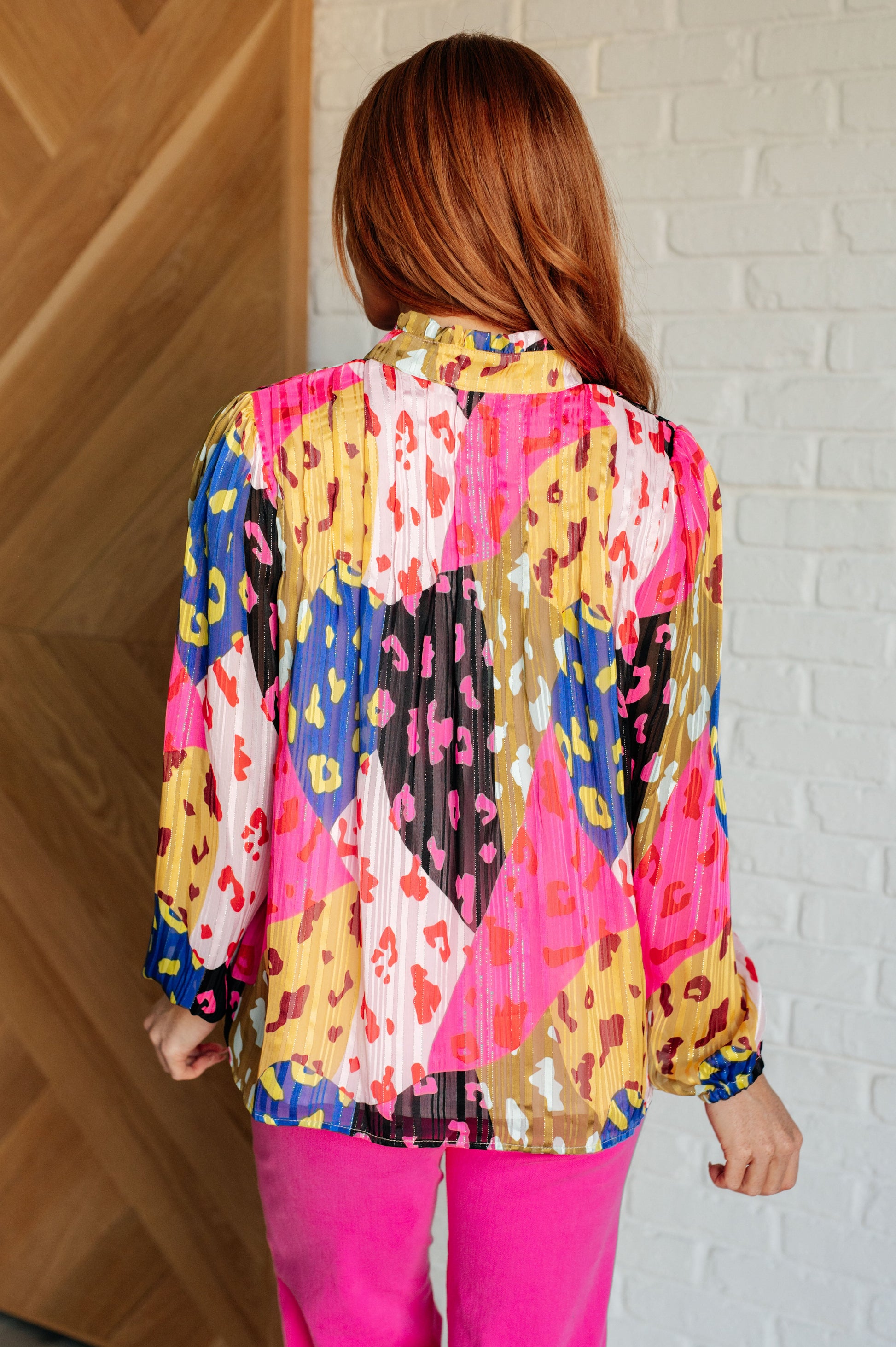 Woman wearing a colorful patterned blouse with pink pants against a white brick wall.