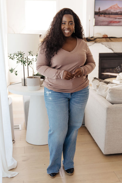 Woman standing in a living room wearing a brown sweater and blue jeans.
