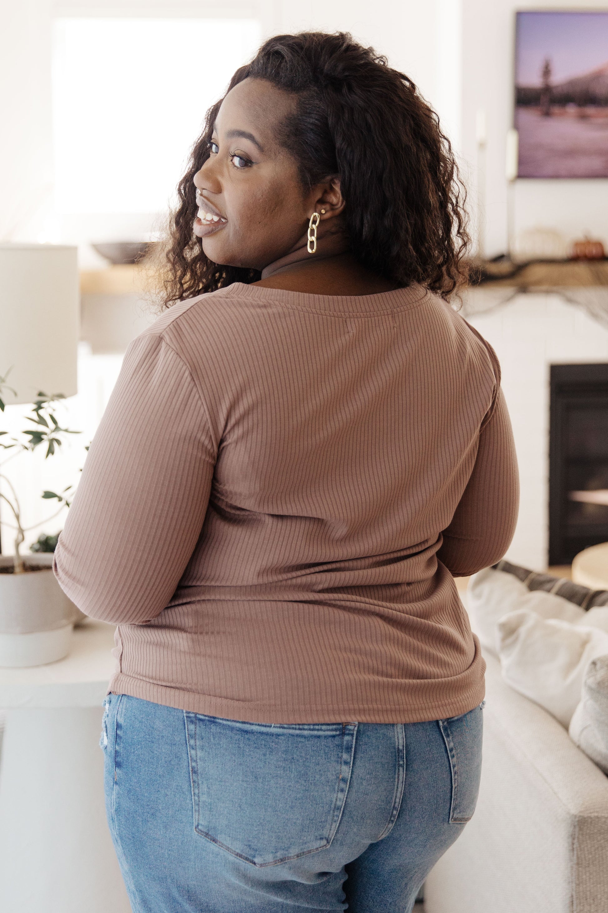 Woman wearing a brown long-sleeve top and blue jeans in a living room setting.