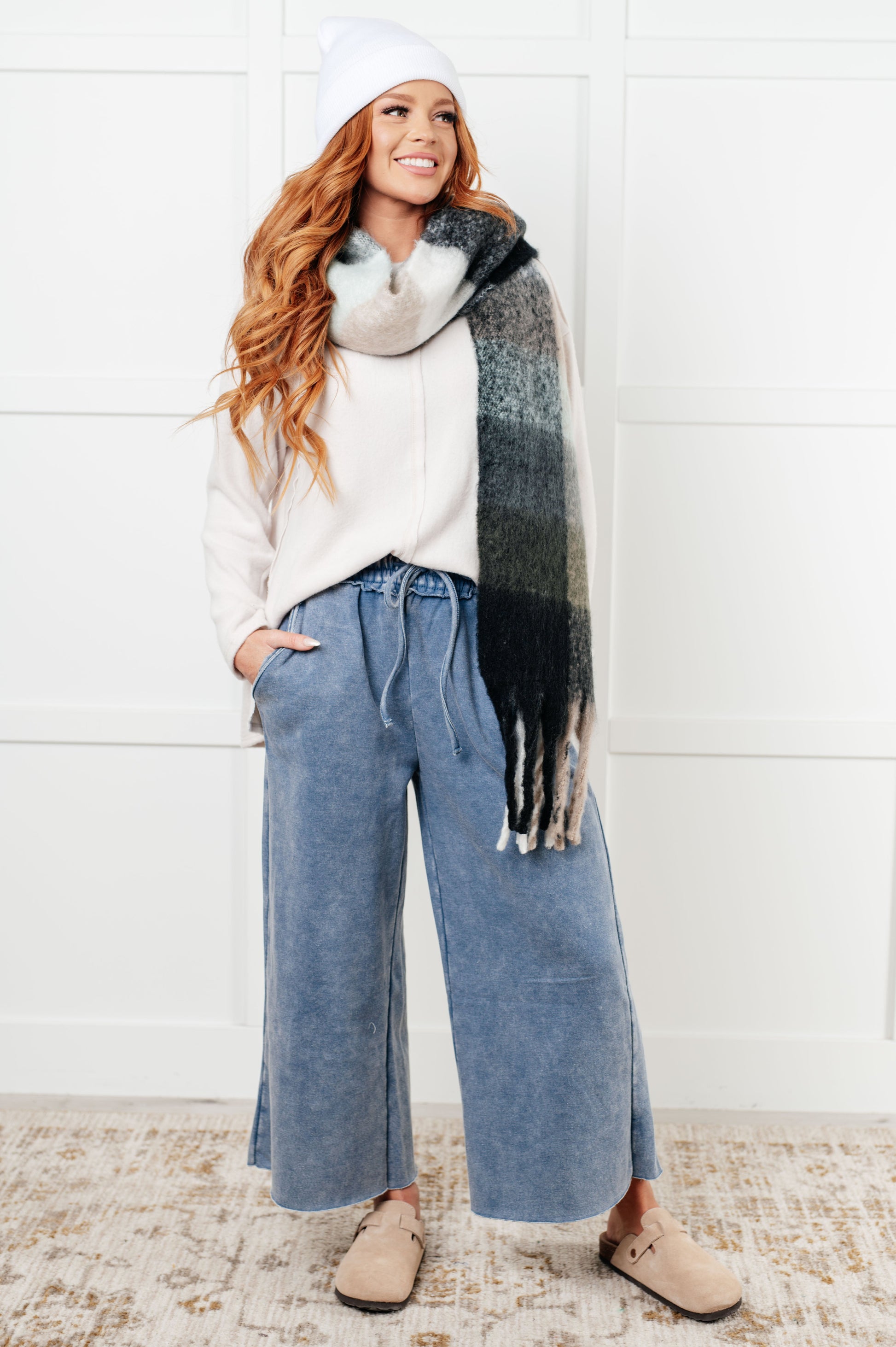 Woman wearing a white sweater, blue jeans, and a patterned scarf against a white wall.