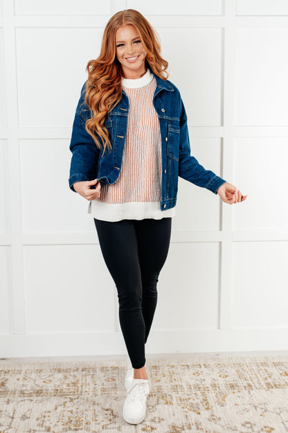 Woman wearing a denim jacket, pink sweater, black leggings, and white sneakers in a room with a white wall.