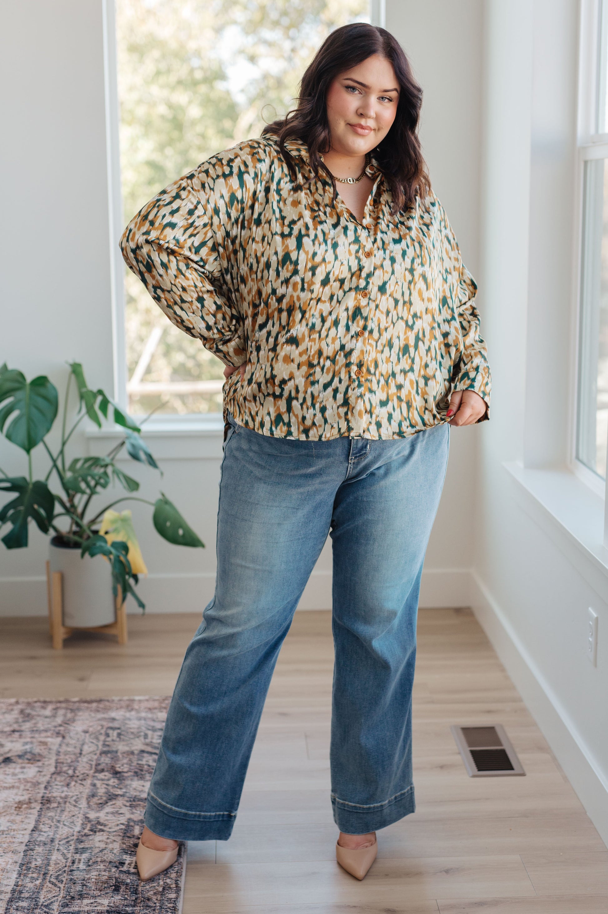 Woman wearing a patterned blouse and jeans standing in a room with a plant and window.