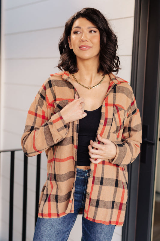 Woman wearing a plaid shirt over a black top and jeans, standing against a white wall.
