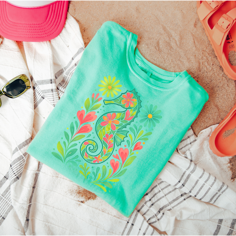 Turquoise t-shirt with floral seahorse design on a sandy surface with beach accessories.