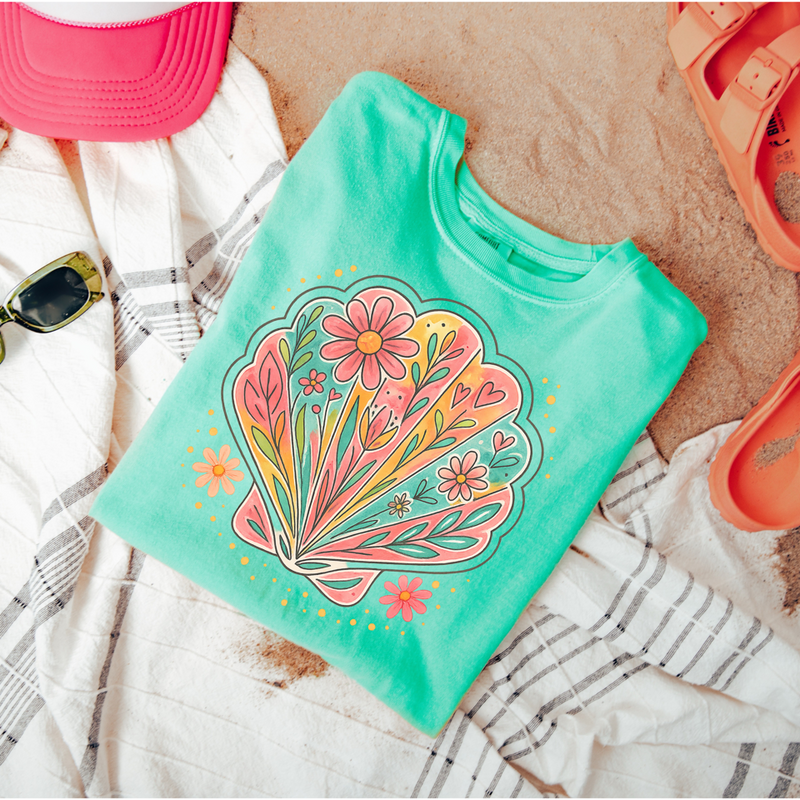 Green t-shirt with floral design on a sandy surface with beach accessories.