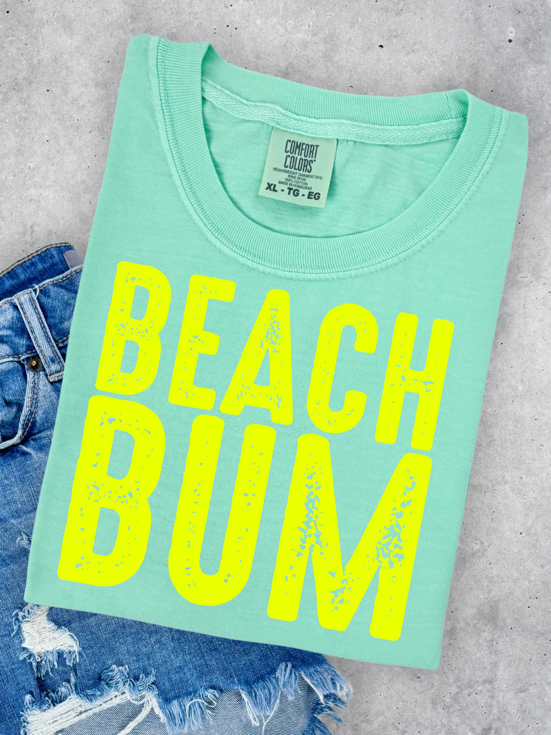 Mint green t-shirt with 'BEACH BUM' in yellow on a concrete surface with denim jeans.
