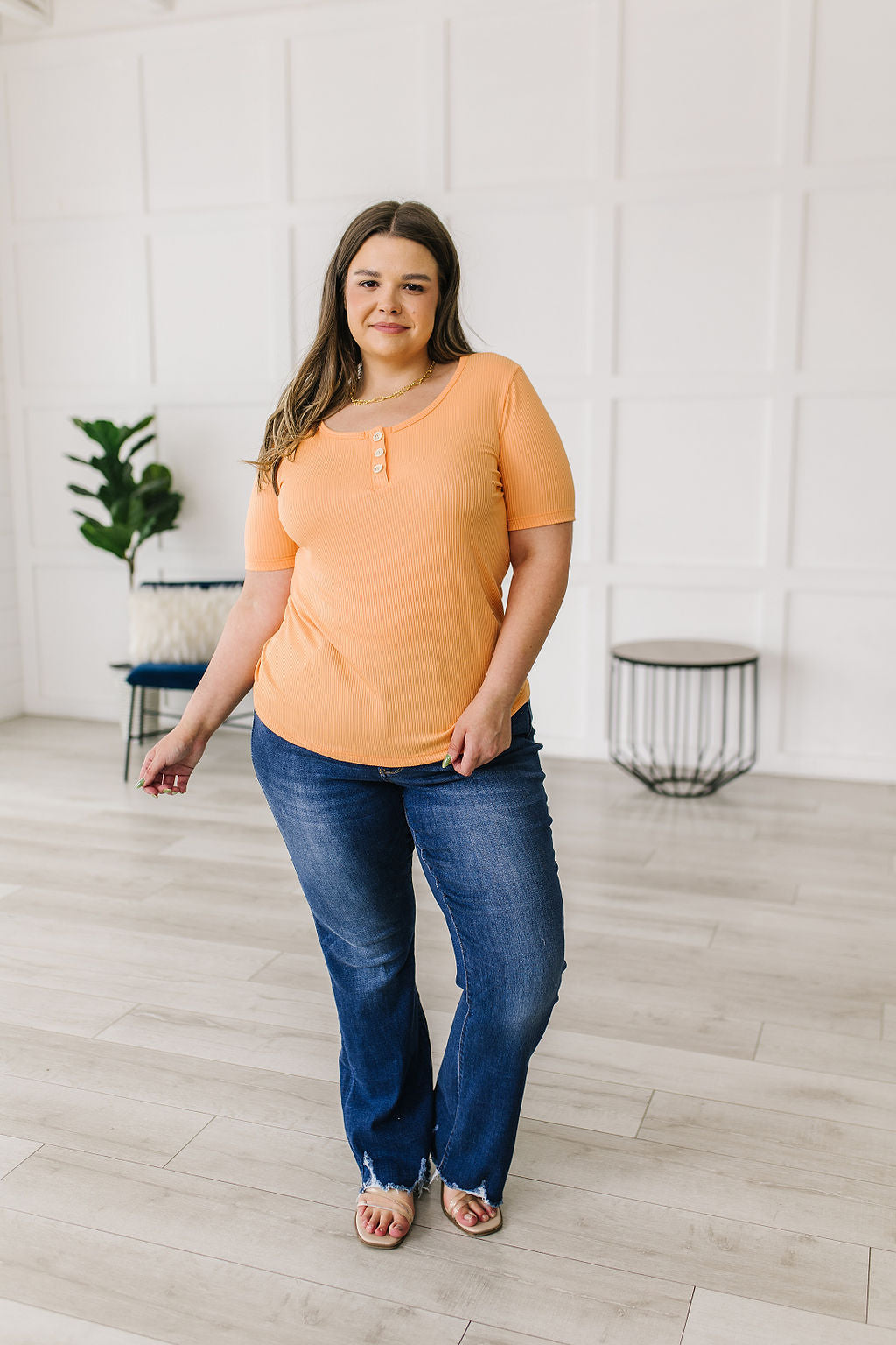 Woman wearing an orange top and blue jeans standing in a room with white walls and wooden flooring.