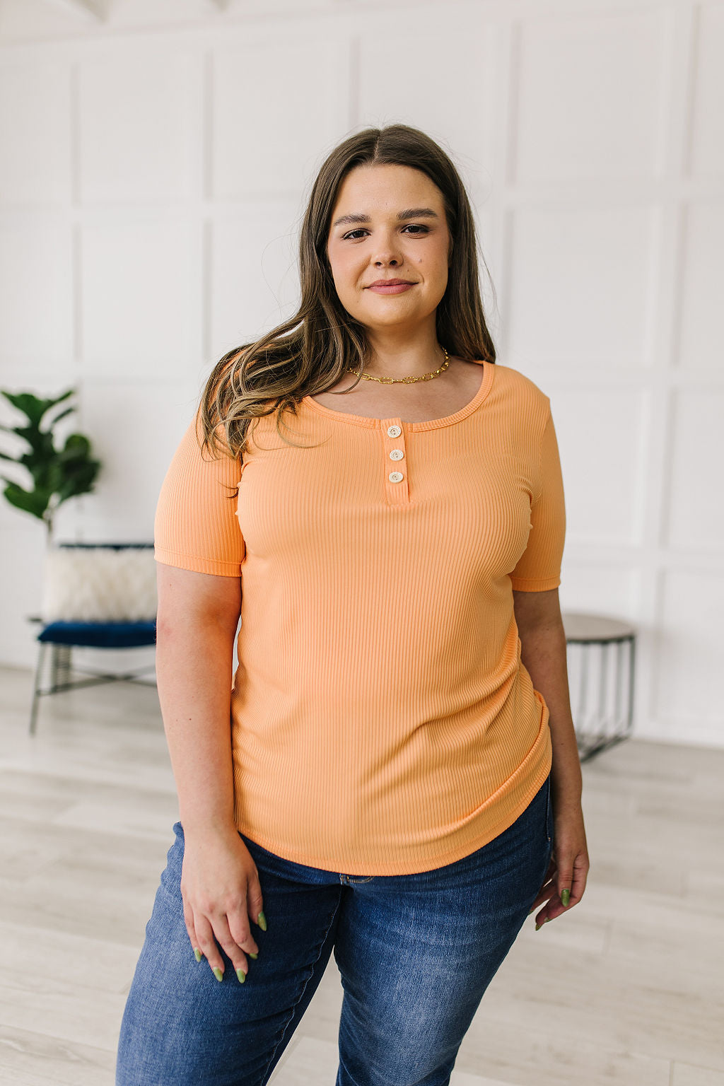 Woman wearing an orange top and blue jeans standing indoors with a white wall background