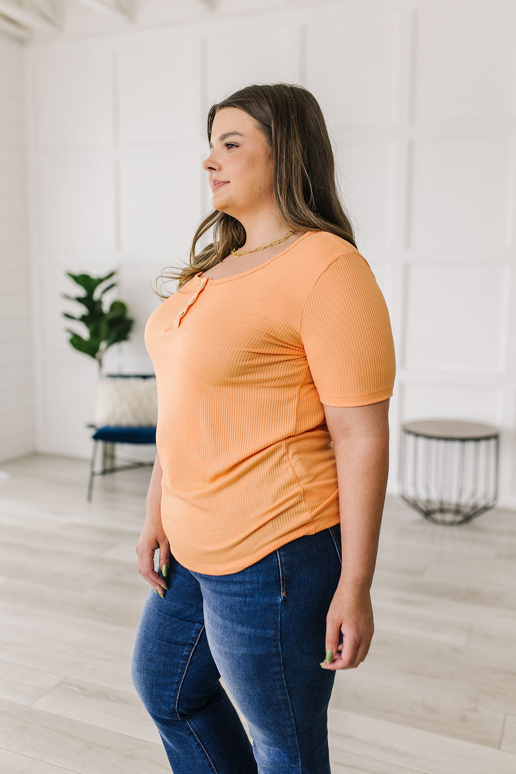Woman wearing an orange top and blue jeans in a bright room with white walls and wooden floor.