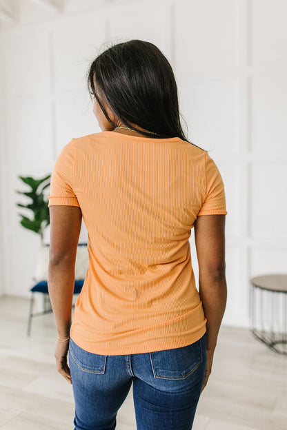 Person wearing an orange ribbed t-shirt and blue jeans standing in a room with a white wall and plant.