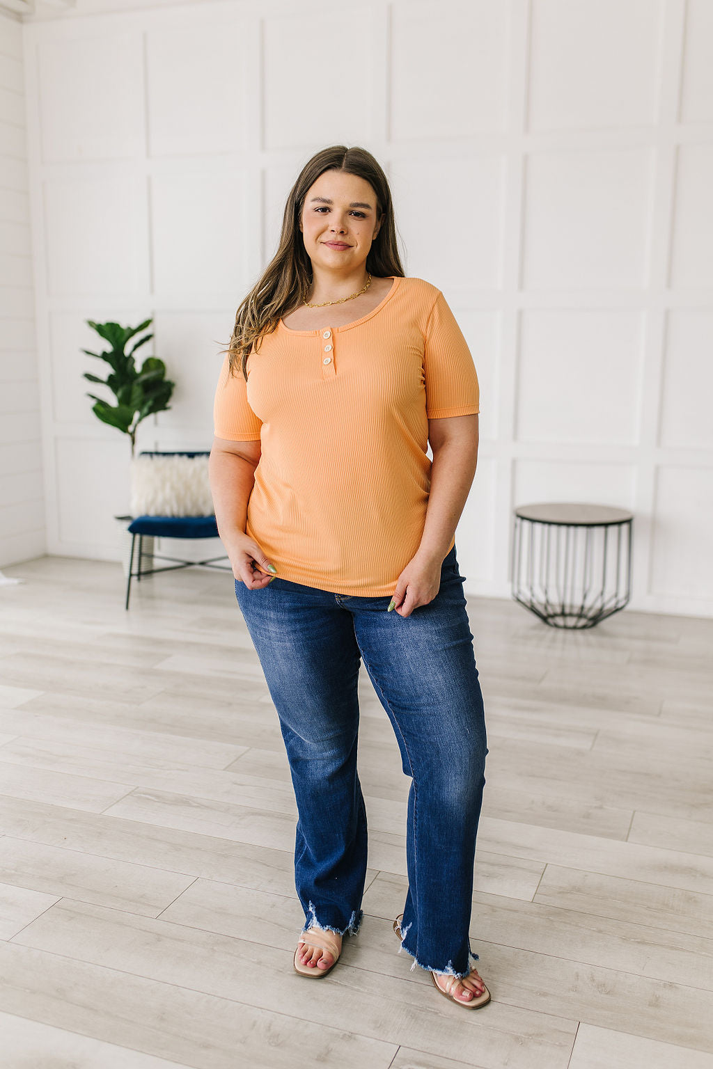 Woman wearing an orange top and blue jeans standing in a room with white walls and wooden flooring.