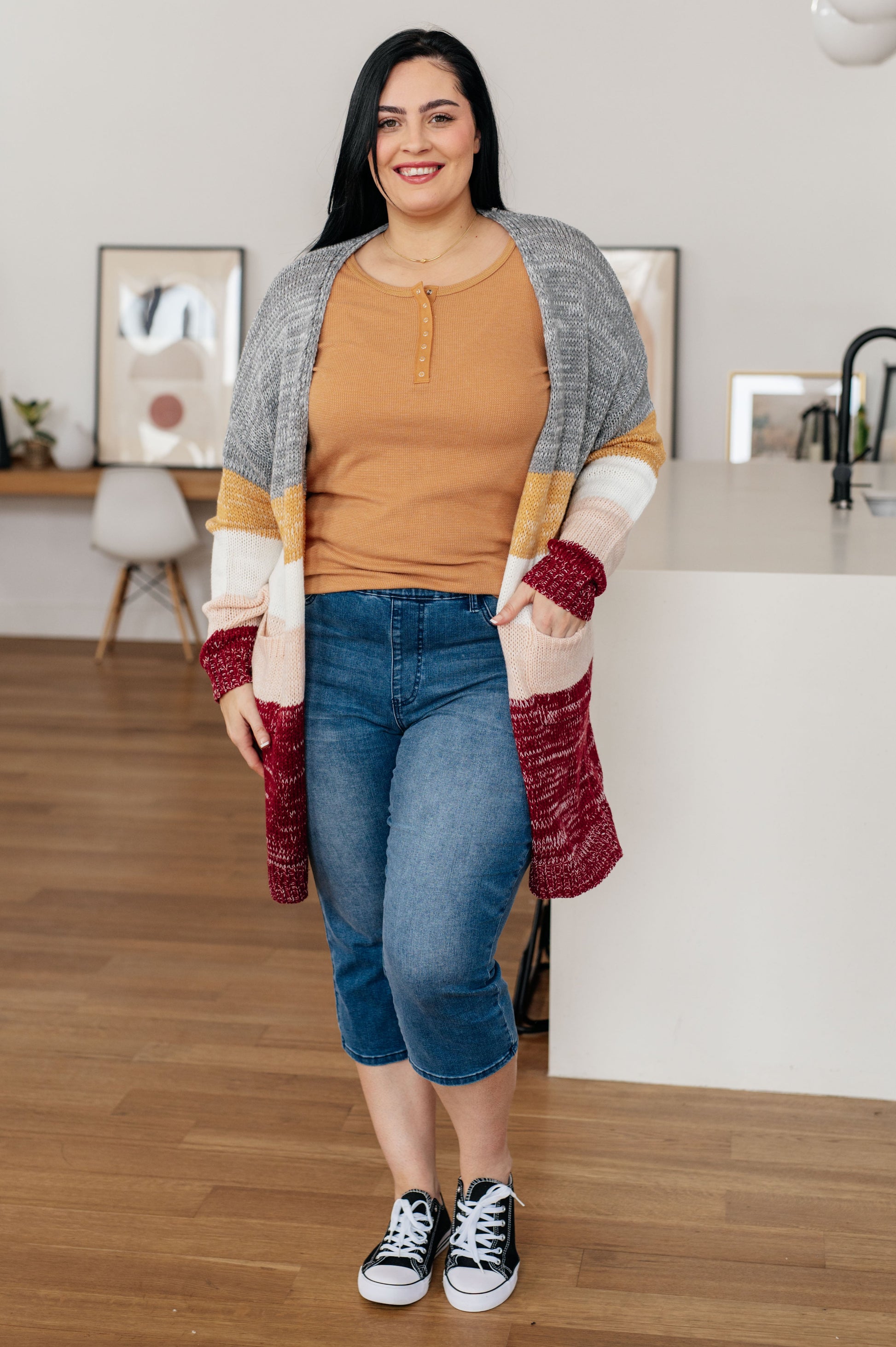 Woman wearing a patterned cardigan, brown shirt, blue jeans, and black sneakers in a kitchen.