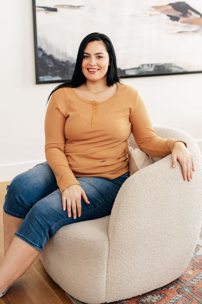 Woman sitting on a beige armchair wearing a mustard yellow top and blue jeans, with a blurred painting in the background.