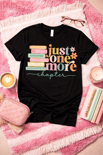 Black t-shirt with colorful text 'just one more chapter' on a pink textured surface with books and glasses.