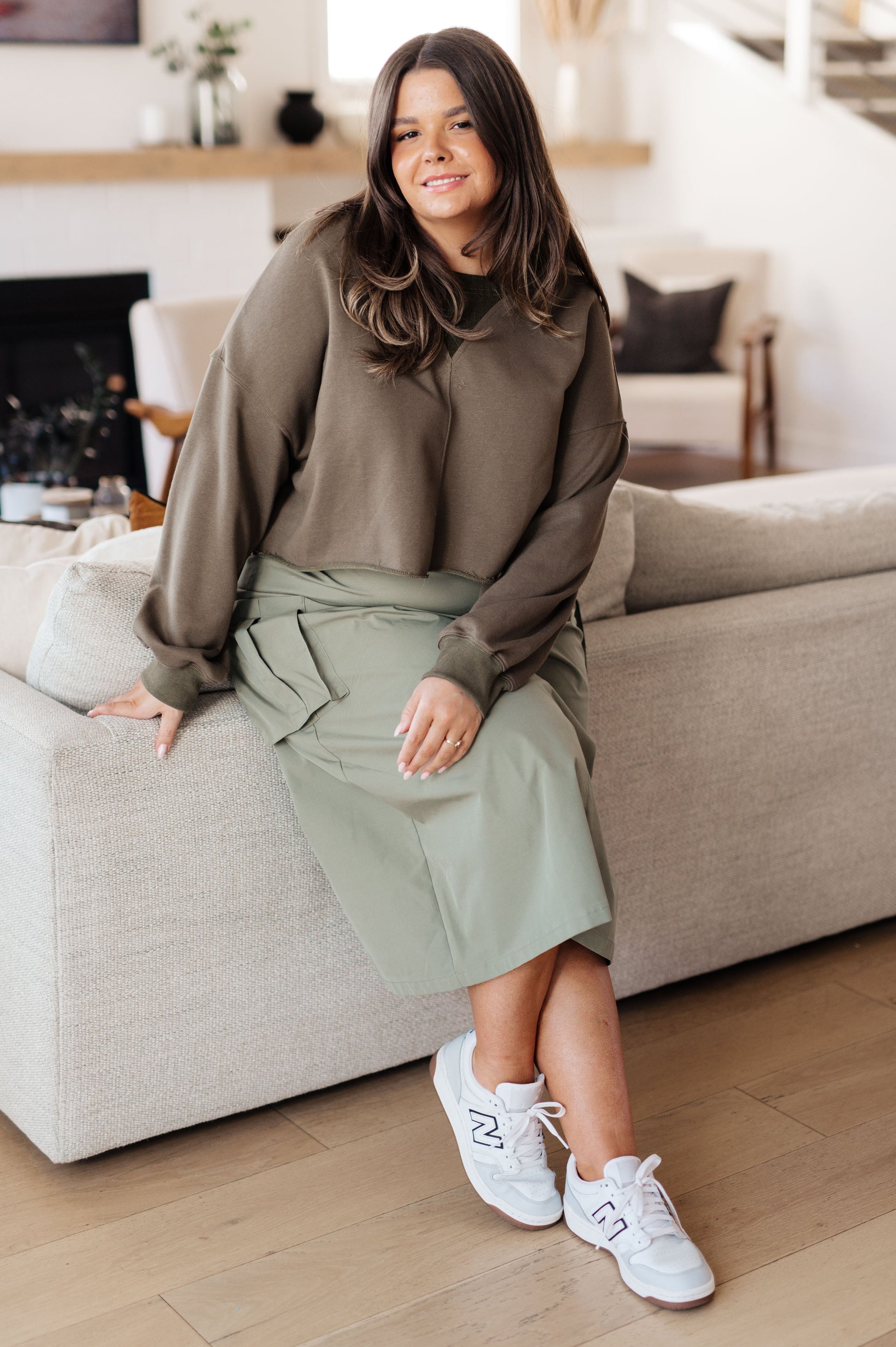 Woman sitting on a couch wearing a brown sweater and green skirt with white sneakers in a living room.