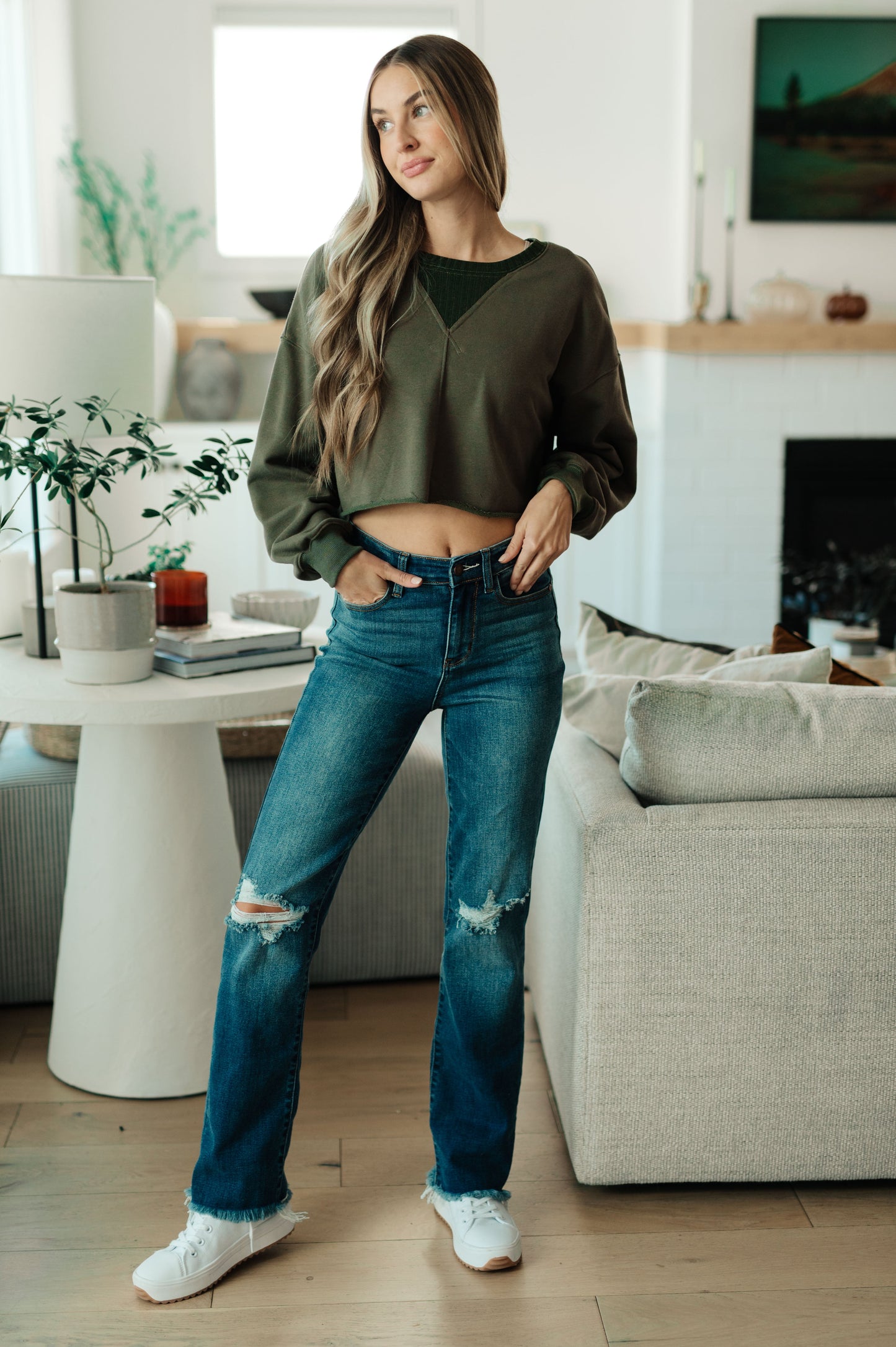 Woman standing in a modern living room wearing an olive green sweater and blue jeans.