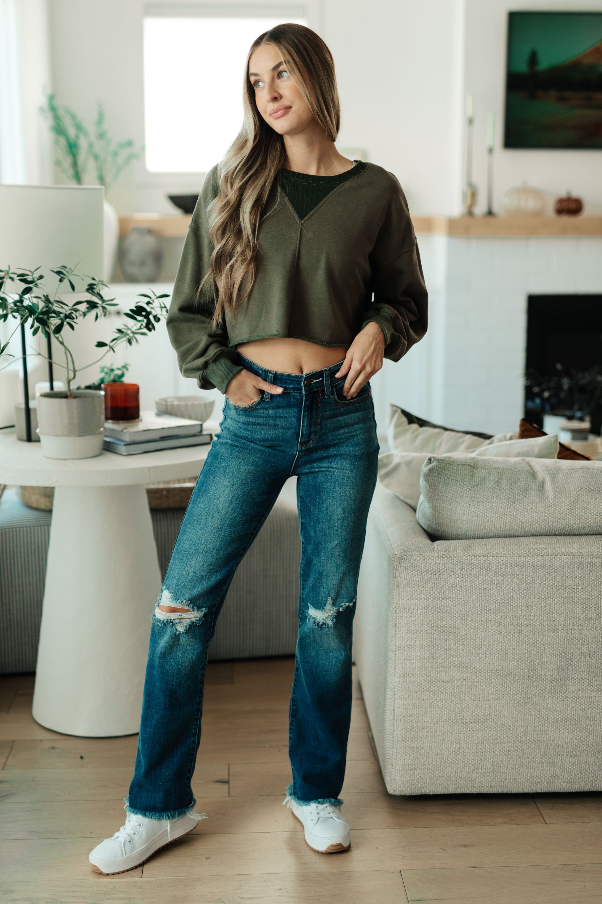 Woman standing in a modern living room wearing an olive green sweater and blue jeans.