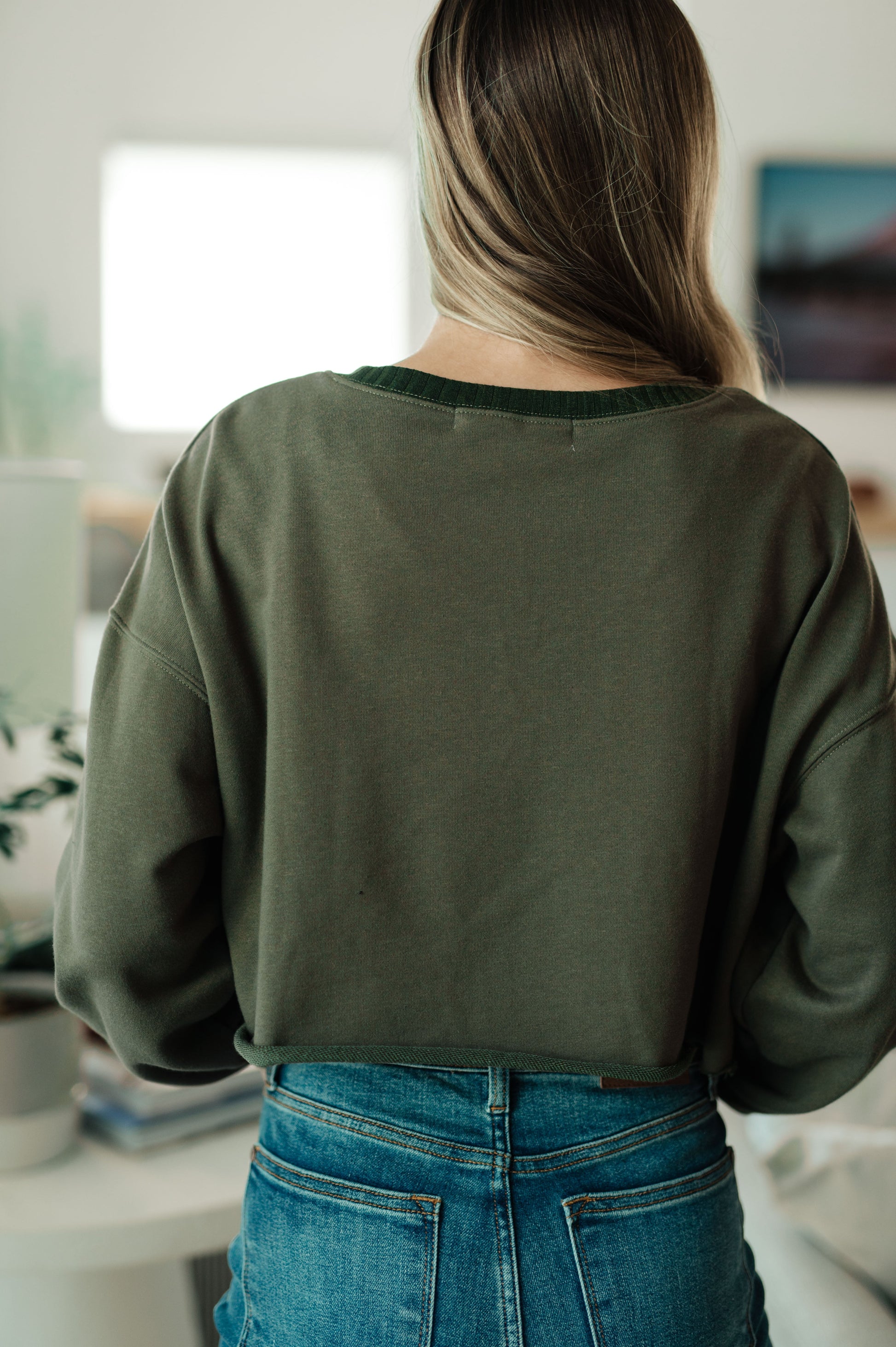 Person wearing an olive green sweatshirt and blue jeans in a room with a blurred background