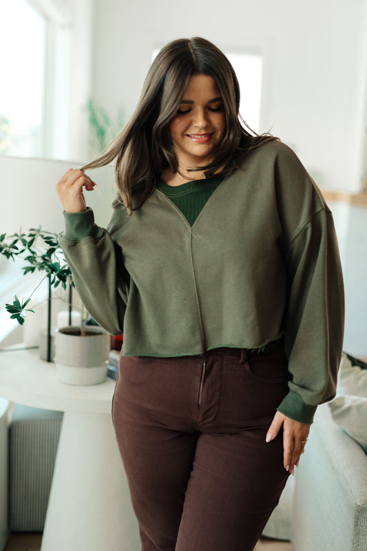 Woman wearing a green sweater and brown pants in a bright room with plants.