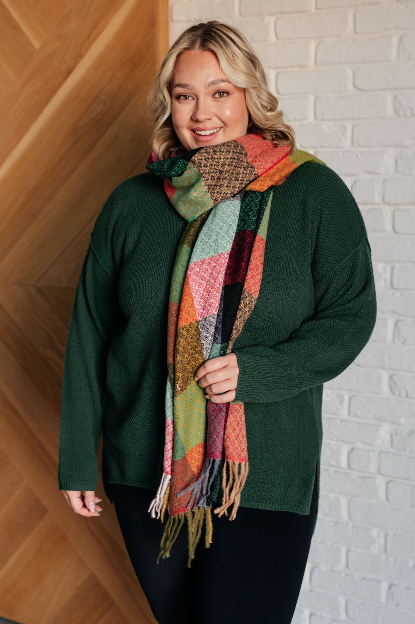 Woman wearing a green sweater and colorful patchwork scarf against a white brick wall.
