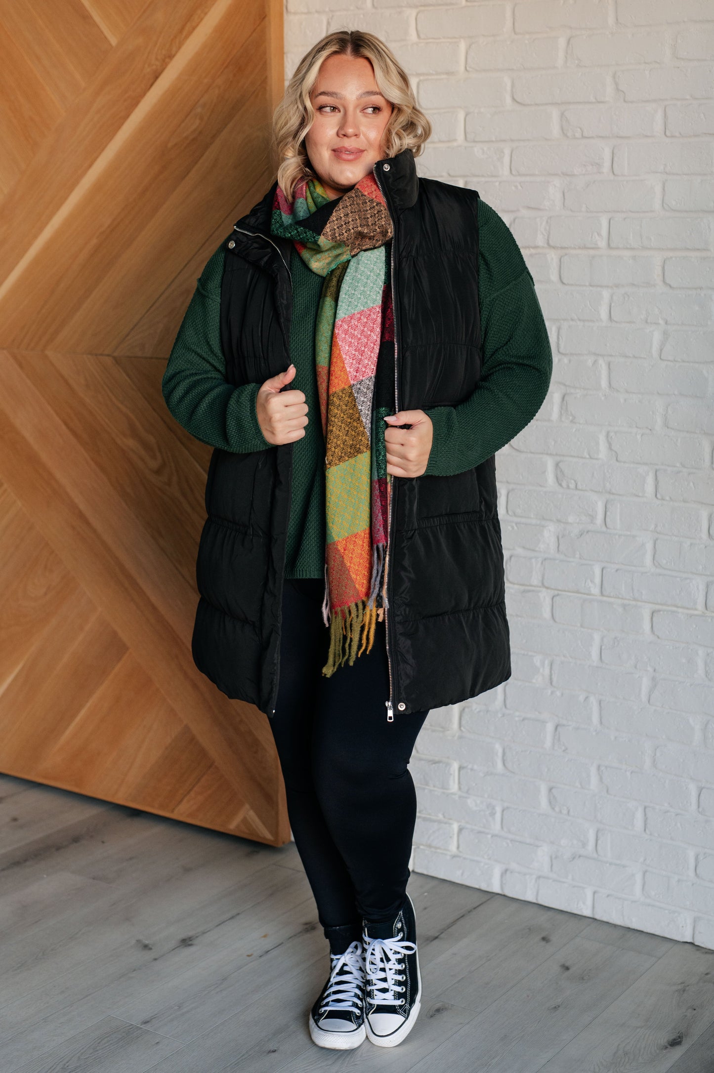 Woman wearing a black coat, green sweater, colorful scarf, and black pants against a white brick wall.