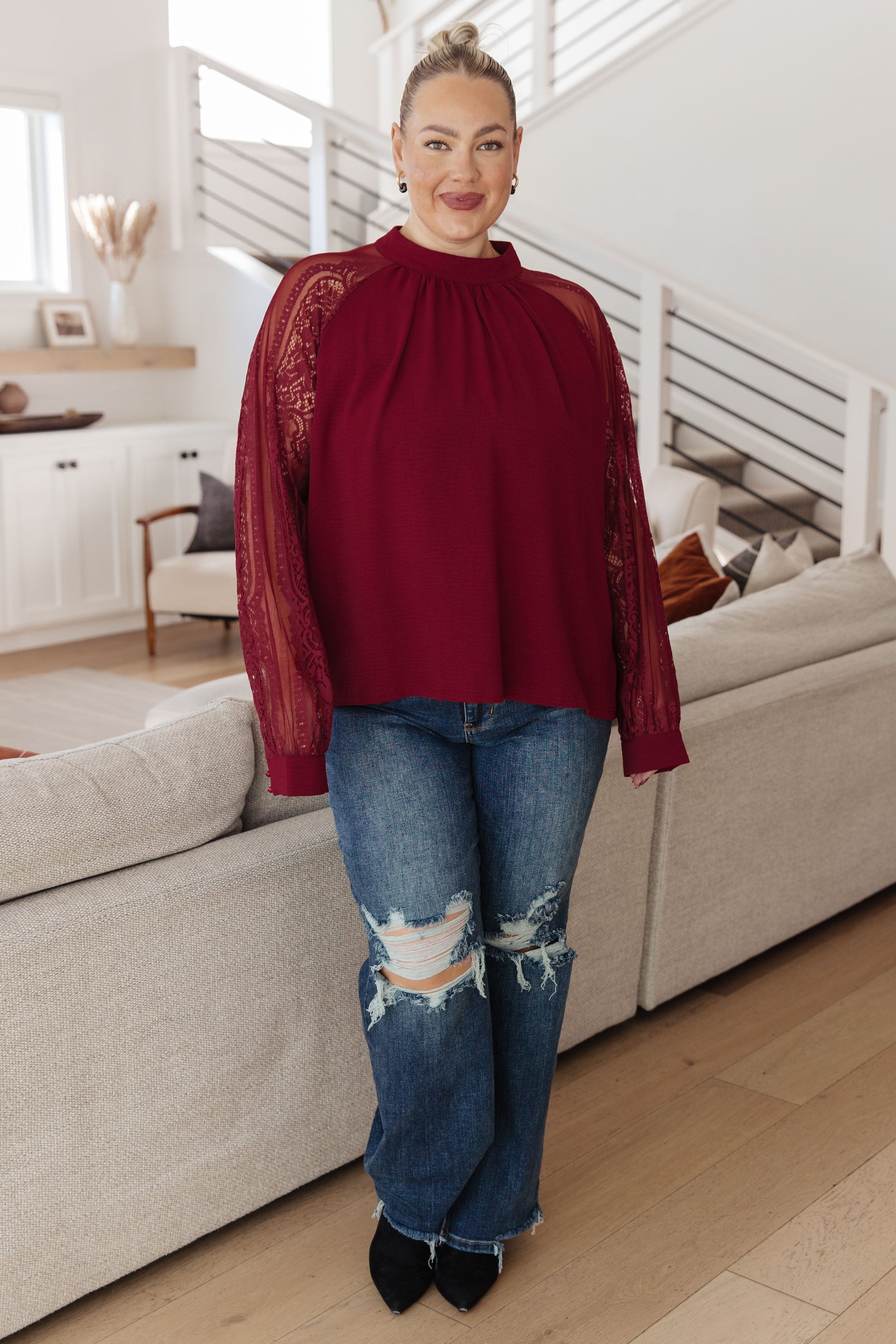 Woman wearing a red blouse with lace sleeves and ripped jeans in a living room.