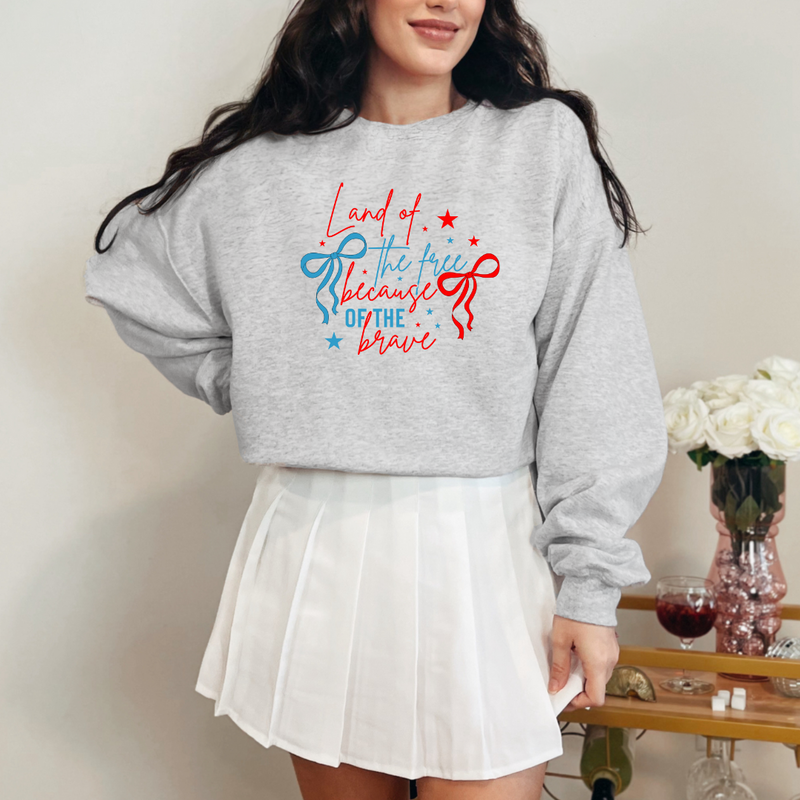 A woman wearing a gray crewneck sweatshirt with the text 'Land of the Free, Because of the Brave' printed on the front.
