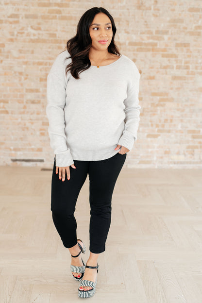 Woman wearing a light gray sweater and black pants against a brick wall.