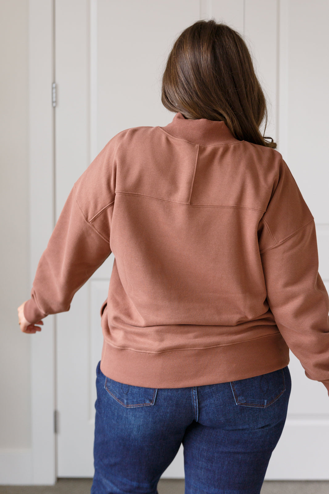 Person wearing a brown jacket and blue jeans against a white wall