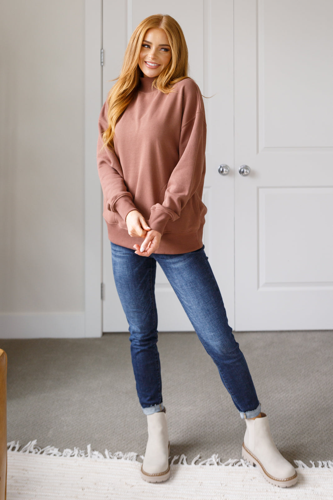 Woman wearing a pink sweater, blue jeans, and white boots standing in a room with a neutral color scheme.