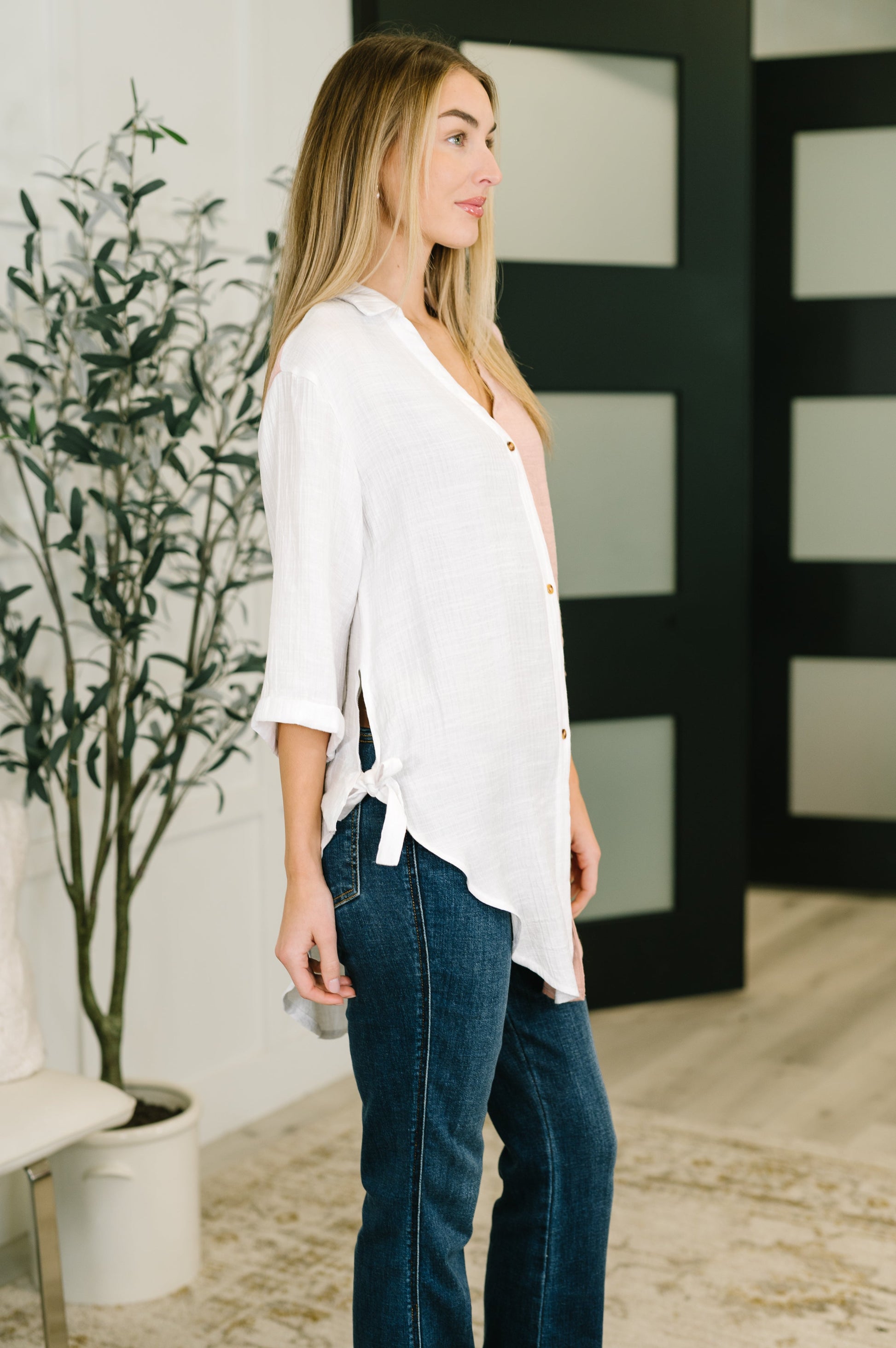 Woman wearing a white blouse and blue jeans standing indoors with a plant and abstract wall art in the background.