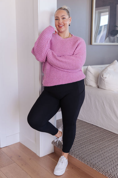 Person wearing a pink sweater and black leggings standing in a room with a couch and framed picture.