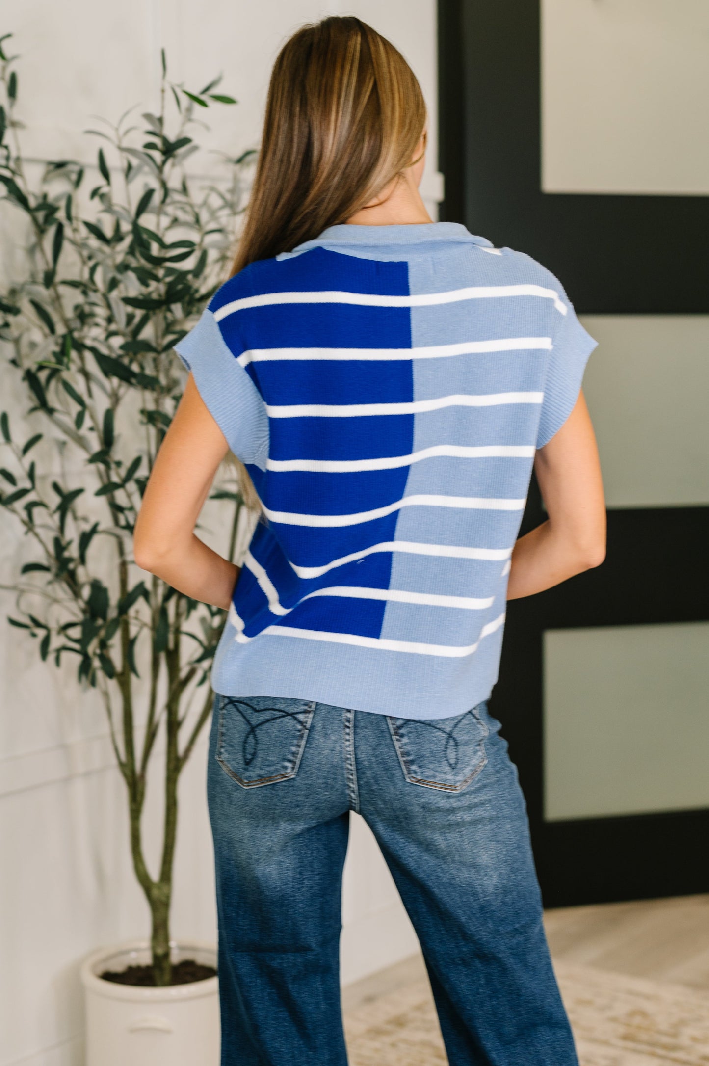 Person wearing a blue and white striped shirt and jeans, standing in a room with a plant and framed picture.