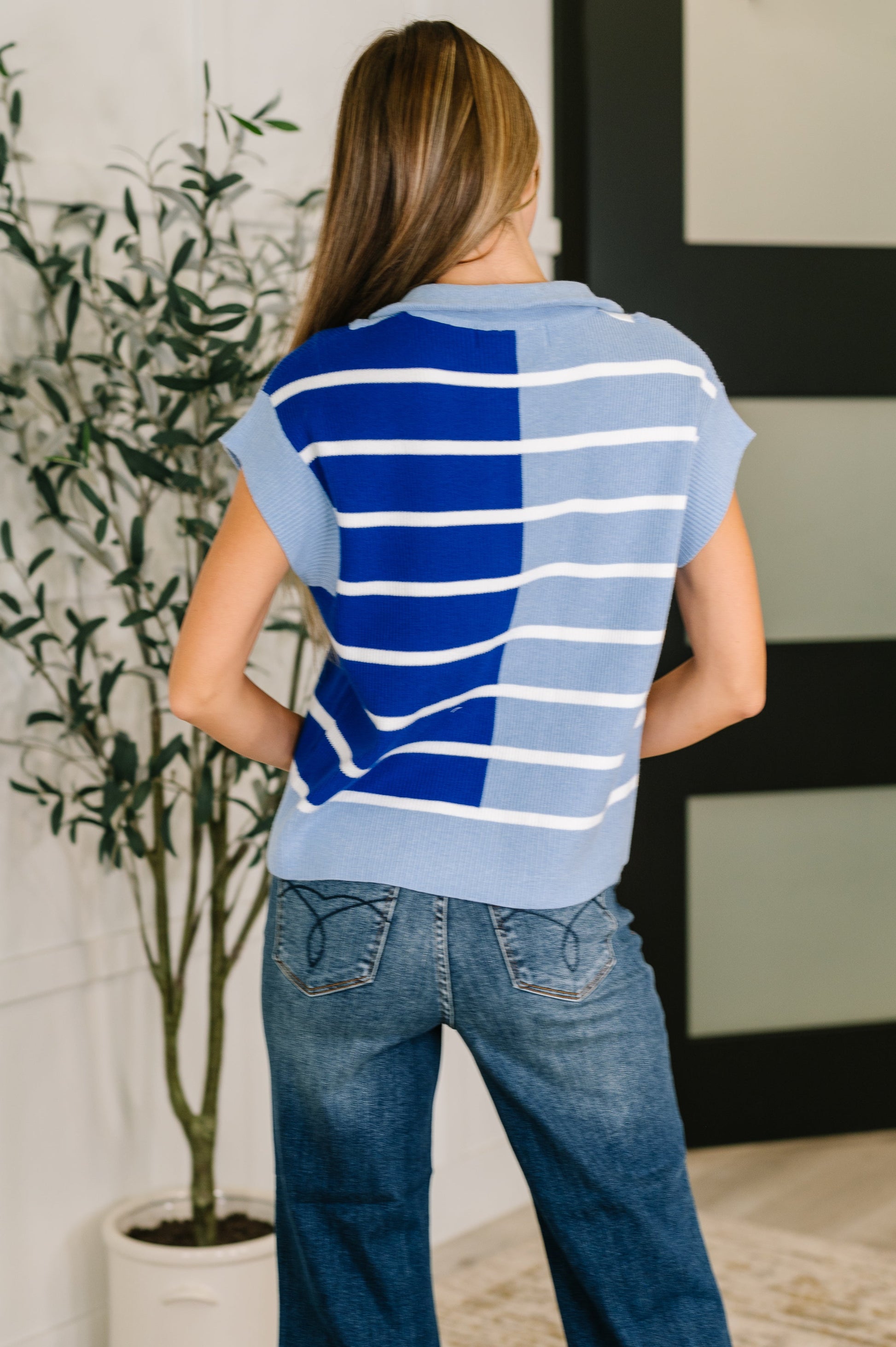 Person wearing a blue and white striped shirt and jeans, standing in a room with a plant and framed picture.