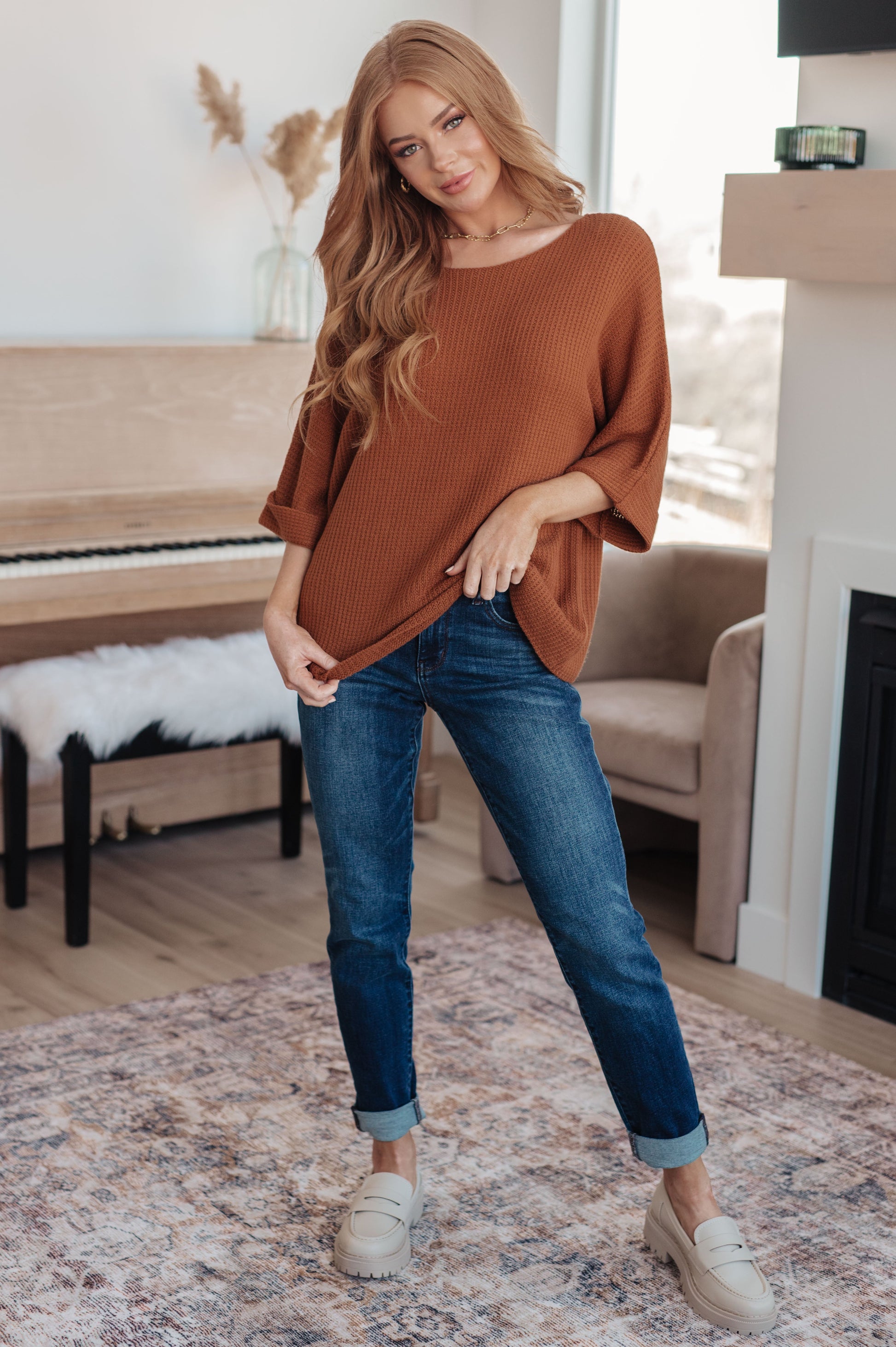Woman wearing a brown sweater and blue jeans standing in a living room.