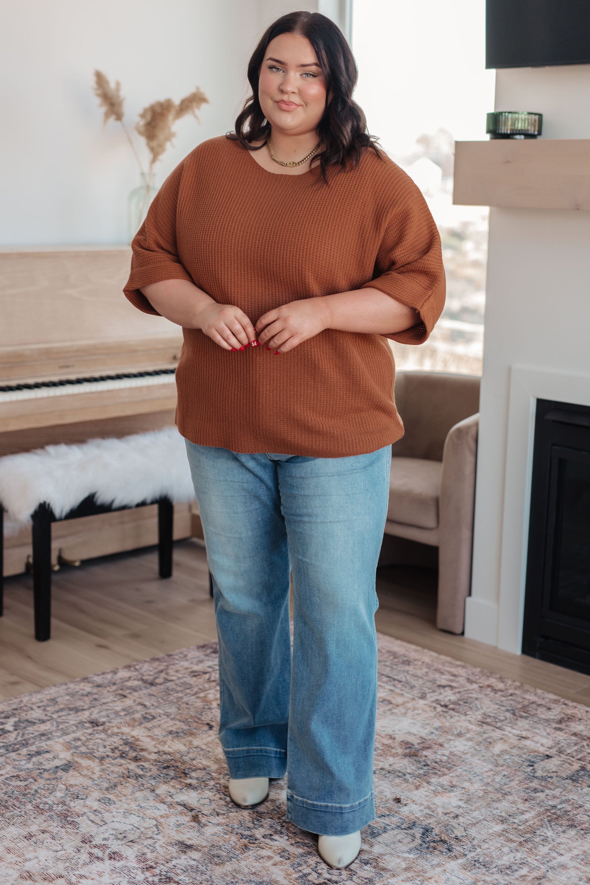 Woman wearing a brown sweater and blue jeans standing in a living room.
